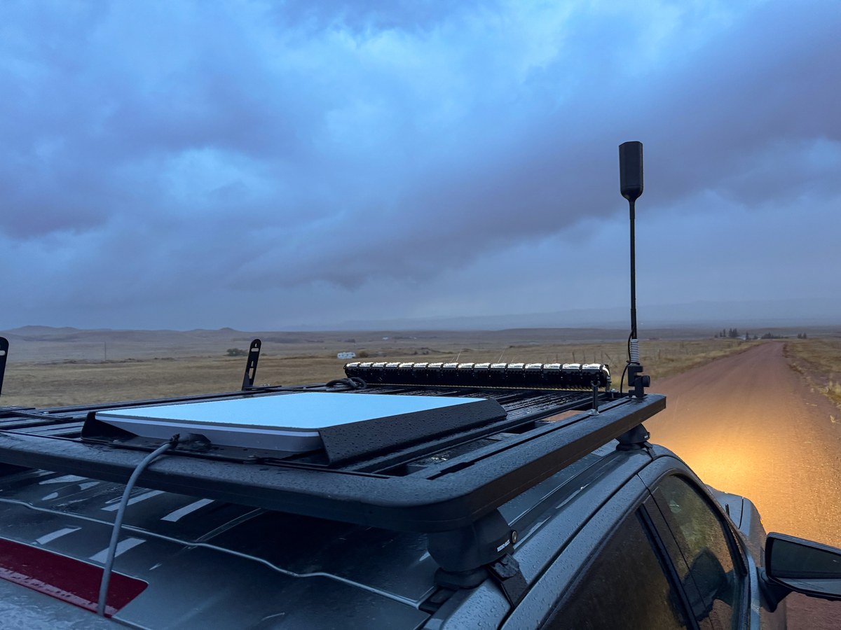 Starlink dish mounted on a pickup truck's roof rack beside a gravel road across open prairie under a cloudy sky